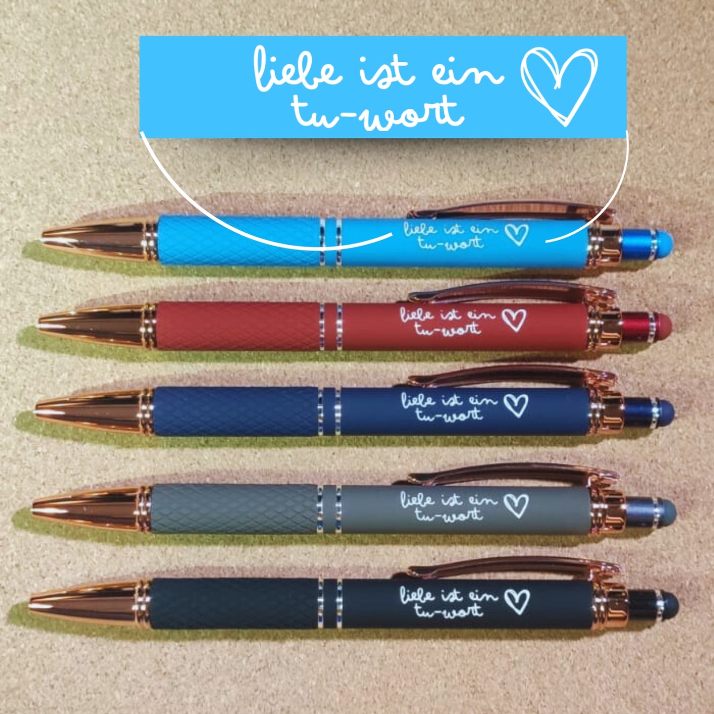 Übersicht der Kugelschreiber "Liebe ist ein Tu-Wort" in fünf Farben: Bordeaux, Hellblau, Marineblau, Taupe und Schwarz. Mit Rosegold-Akzenten, diamantgemustertem Griff und Soft Touch Finish.