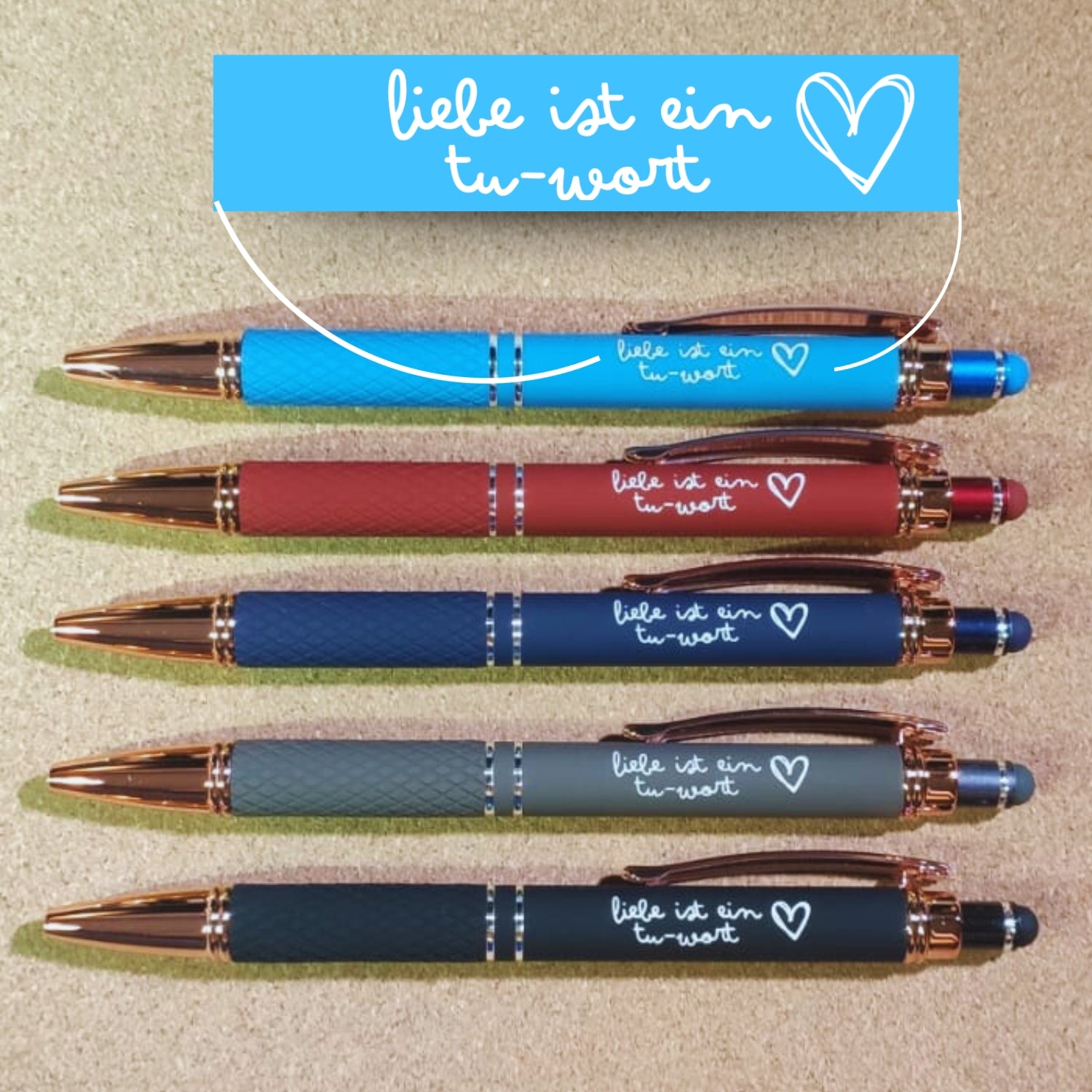 Übersicht der Kugelschreiber "Liebe ist ein Tu-Wort" in fünf Farben: Bordeaux, Hellblau, Marineblau, Taupe und Schwarz. Mit Rosegold-Akzenten, diamantgemustertem Griff und Soft Touch Finish.
